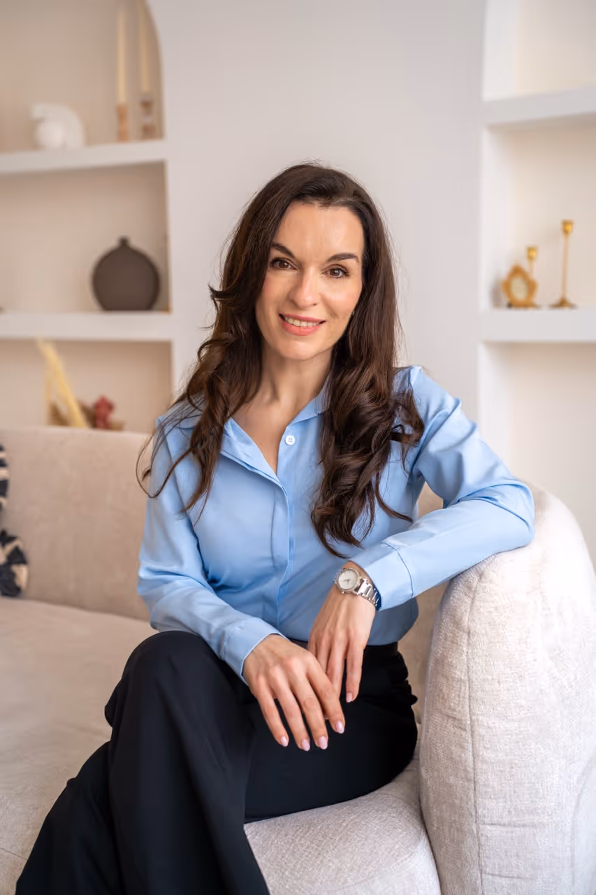 Smiling woman with long dark hair wearing a light blue blouse and black pants seated on a beige couch in a modern living room.