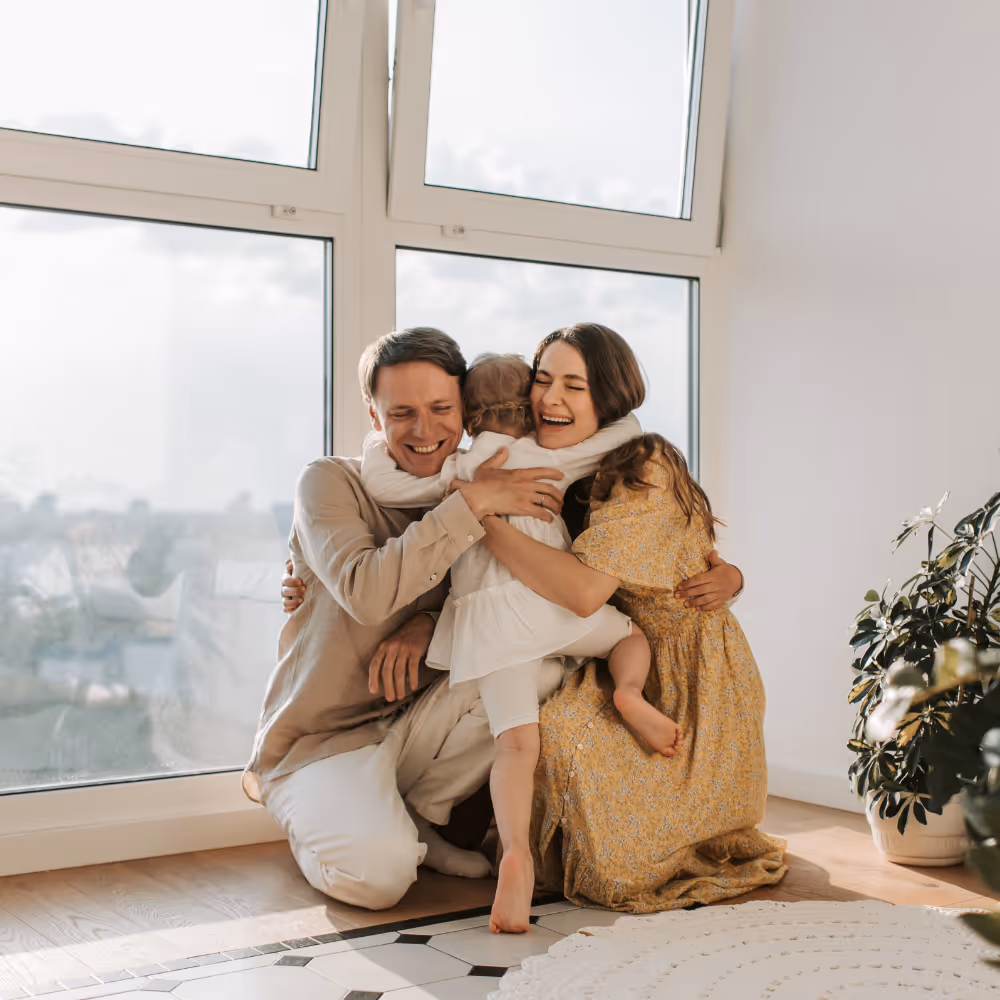 Smiling parents kneeling on floor and hugging their young child in a bright room with large windows and plants.