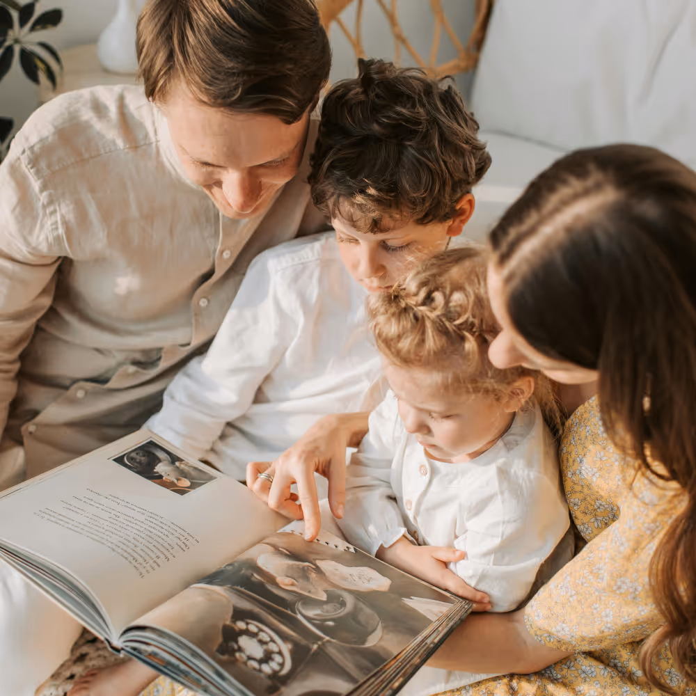 A family of four reading a photo book together, with a mother pointing at a picture.