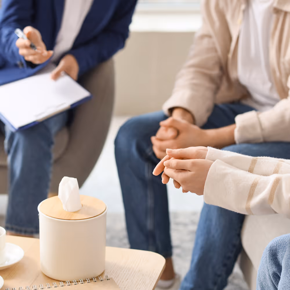 Two people sitting with hands clasped, speaking with a counselor holding a pen and clipboard.